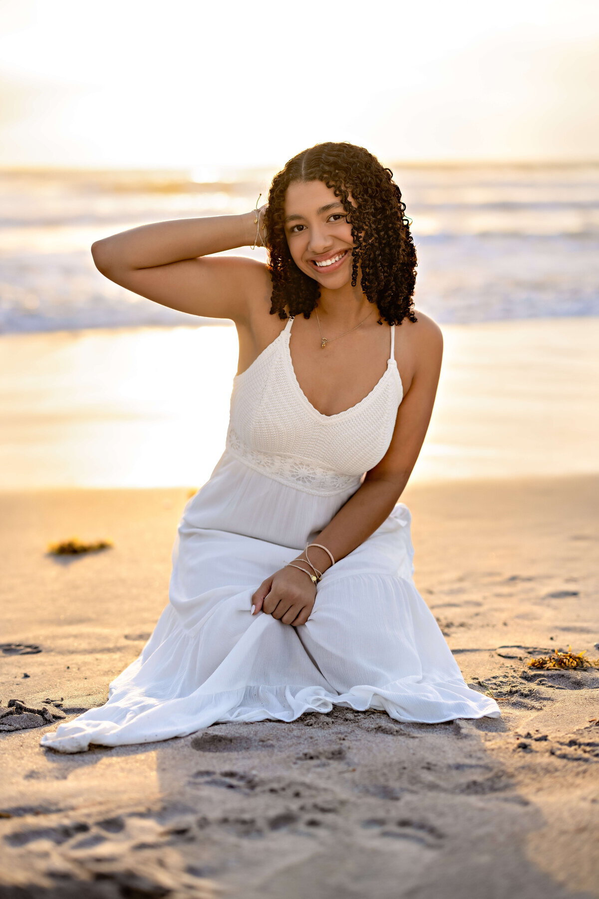 Cocoa Beach senior session with a girl in a white dress, one arm lifted behind her head, smiling in the soft sunrise ight.