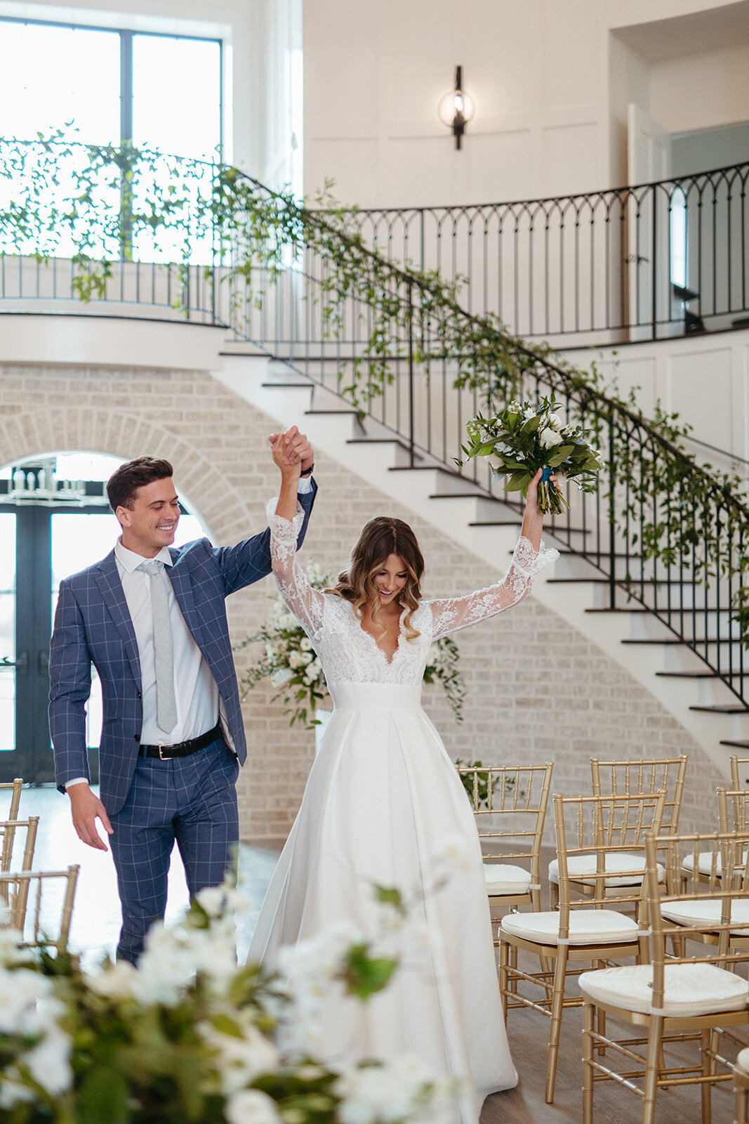 Bride and groom in a blue suit and white wedding gown walking down an aisle of gold chairs with a staircase behind.