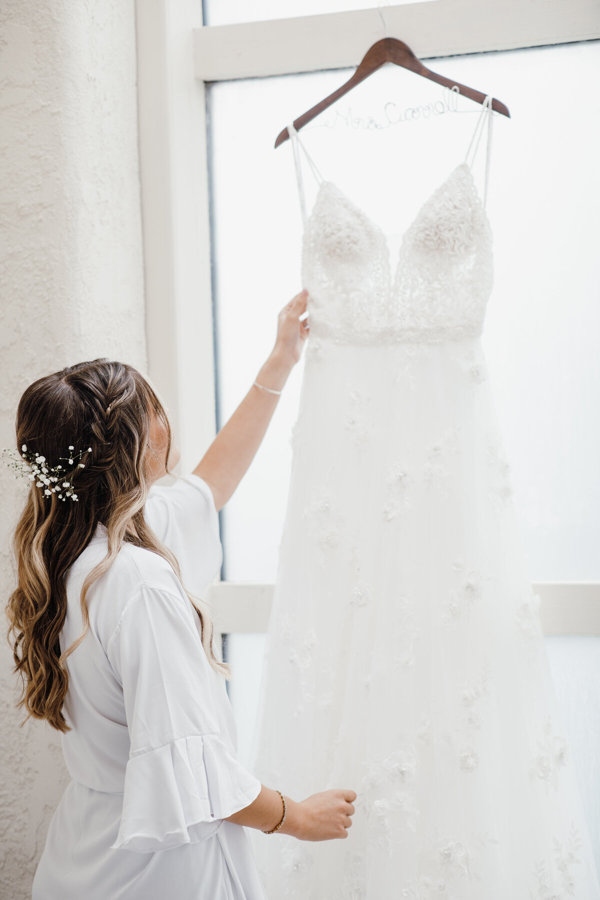 Bride in a white robe touching her wedding dress hanging on a wooden hanger in front of a window