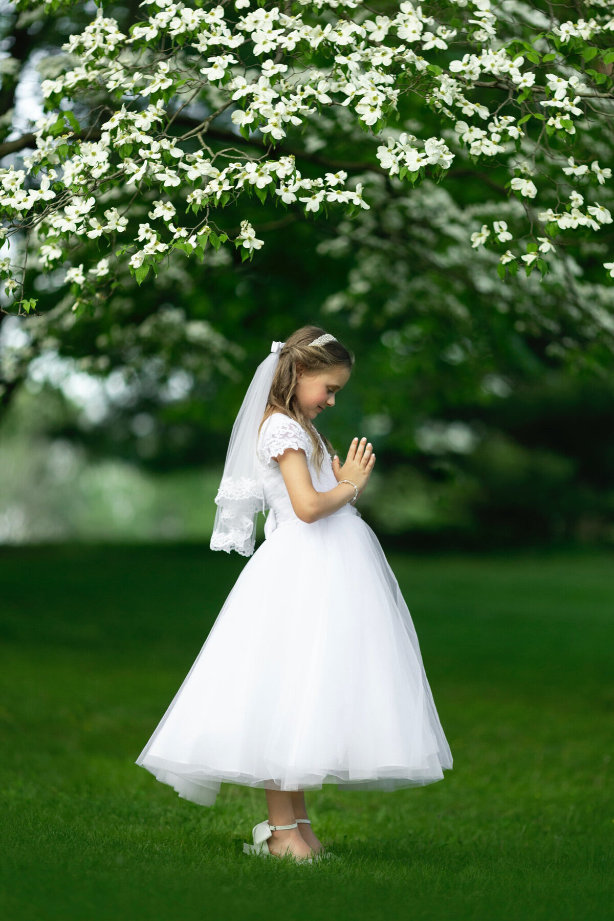A young girl prays in a lawn under some flowering trees in a park wearing a white communion dress