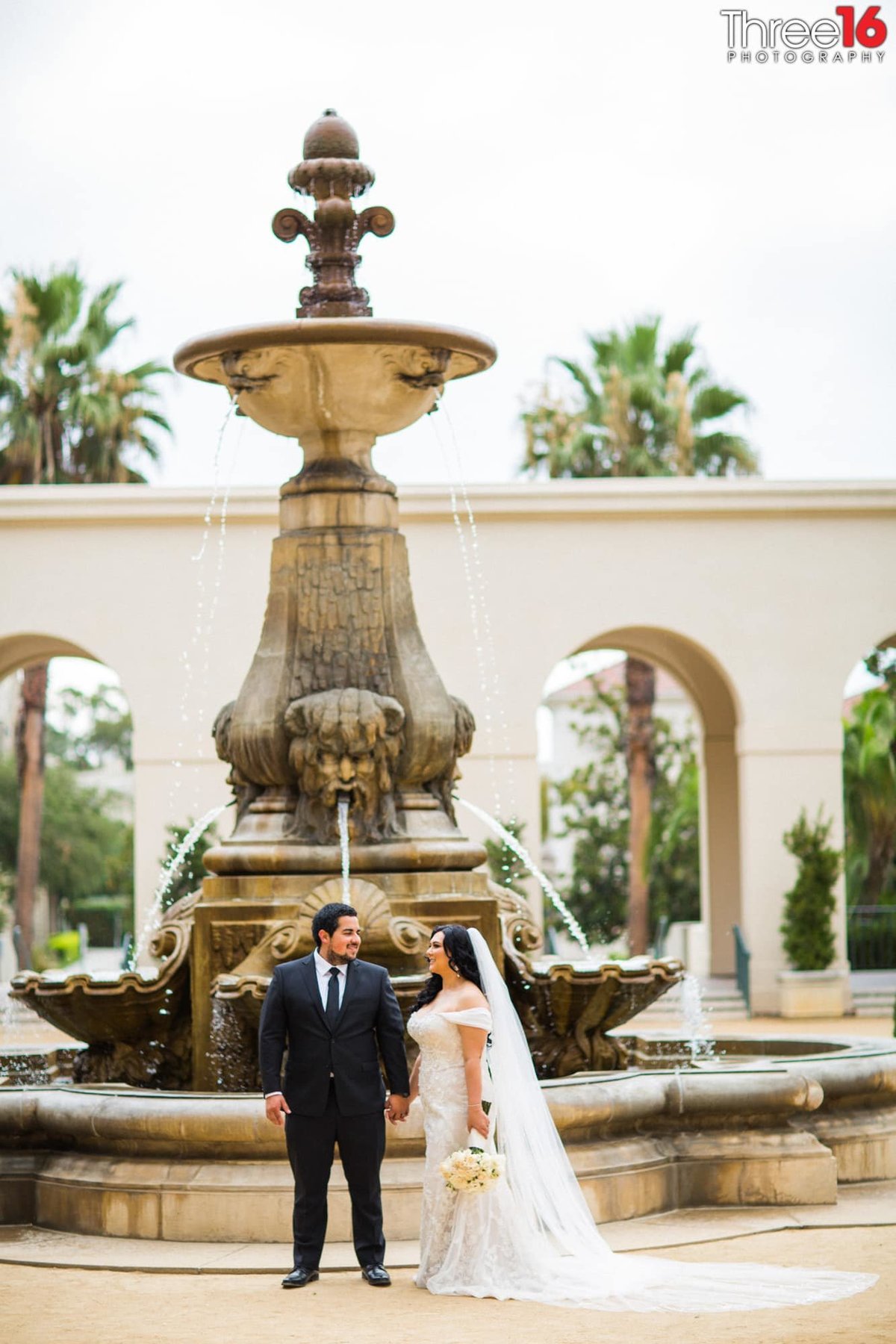 Married couple look at each other while holding hands in front of a large water fountain at the Pasadena City Hall