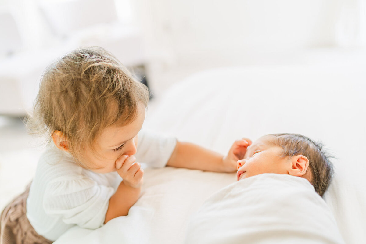 a toddler girl stands next to her newborn brother who is laying on a bed in their Round Rock home and caresses his forehead.