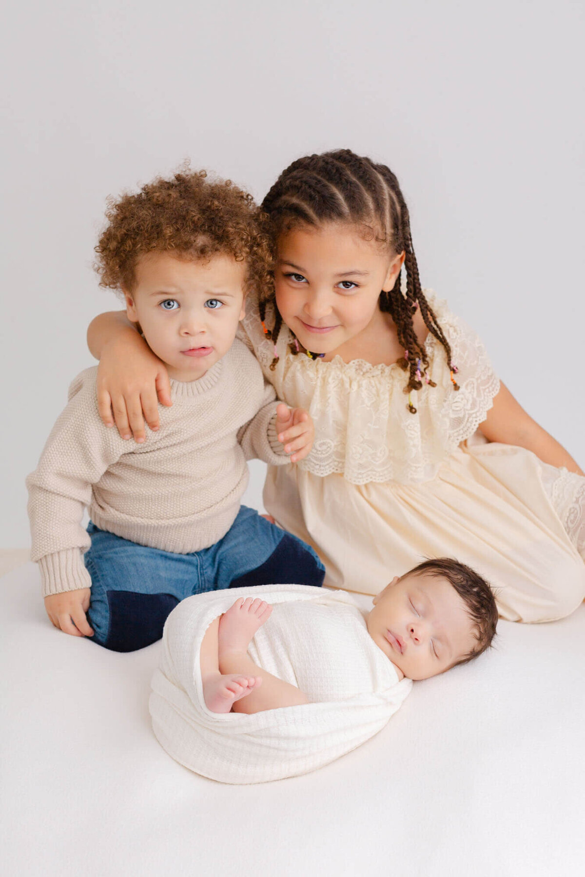Toddler and older sister and close together sitting behind their newborn brother who is swaddled up in white and sleeping during newborn photos.