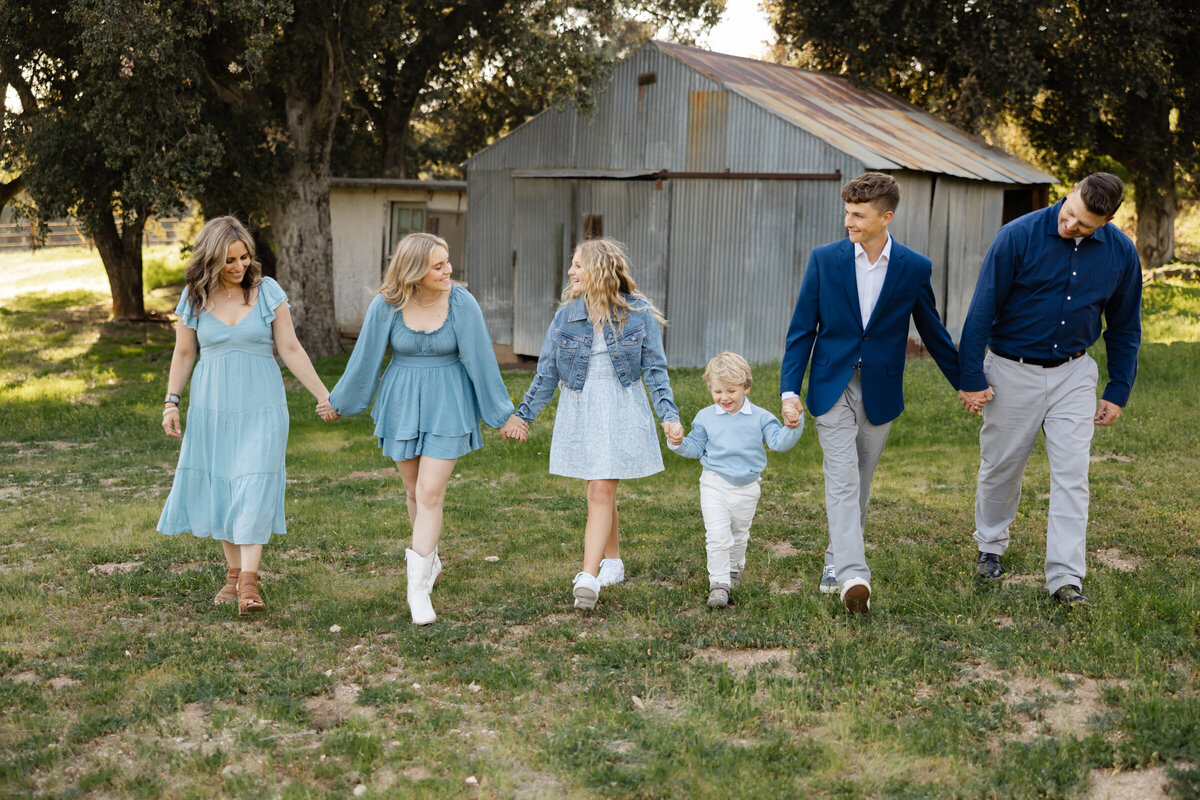 Family of six holding hands and walking together in front of rustic barn, dressed in blue and gray outfits. Photographed by Yucaipa family photographer Kaitlyn Dawn Photography.