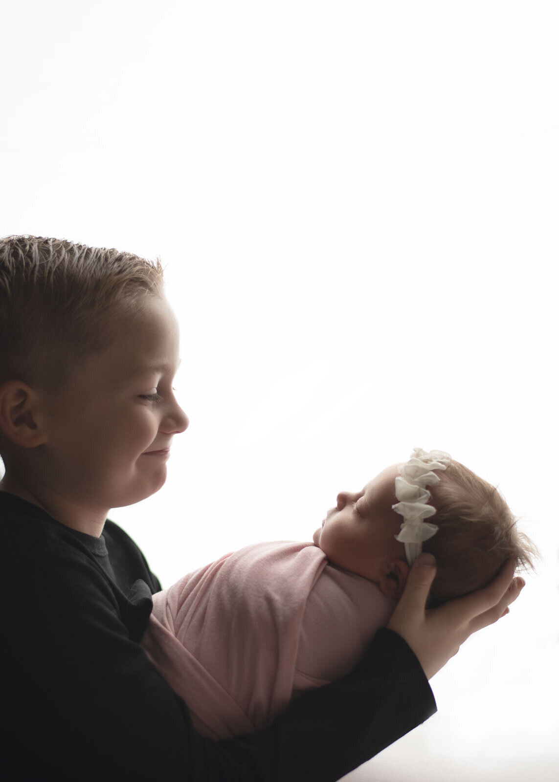 boy holding his baby sister who is wrapped in pink with a white headband. they are on a white background photo taken by sutherland photography, a st. louis newborn photographer