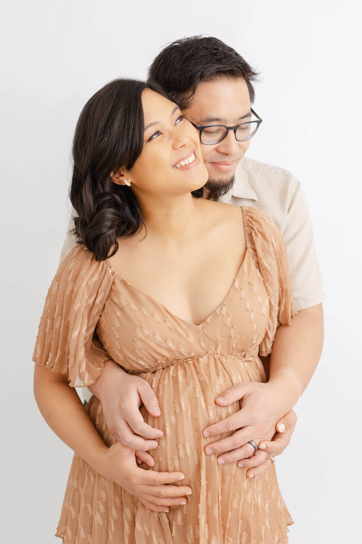 Pregnant woman in a low-cut tan dress holding her belly and looking up towards the light and smiling. Her husband is standing  behind her and has his arms wrapped around her belly. His eyes are closed and he has a small grin on his face. There is a plain white backdrop behind them at an all-white portland maternity portrait studio. Photo by Ashlie Behm Photography