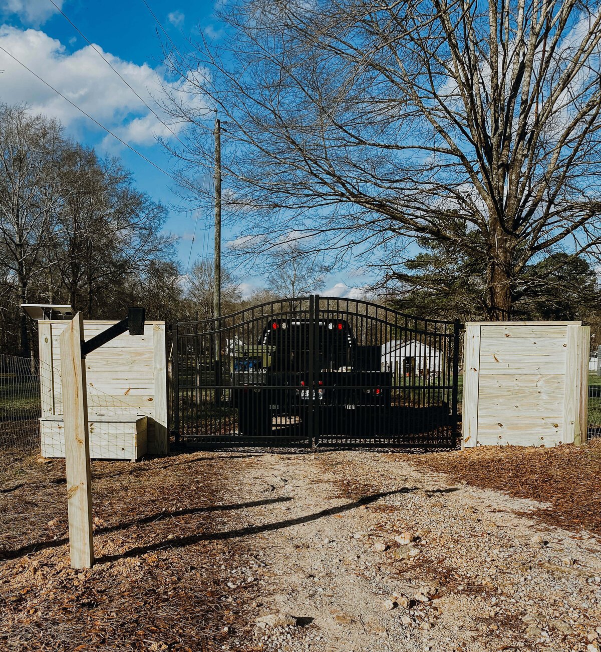 truck-driving-on-gravel-path-through-entrance-gate-under-large-tree-and-blue-skies