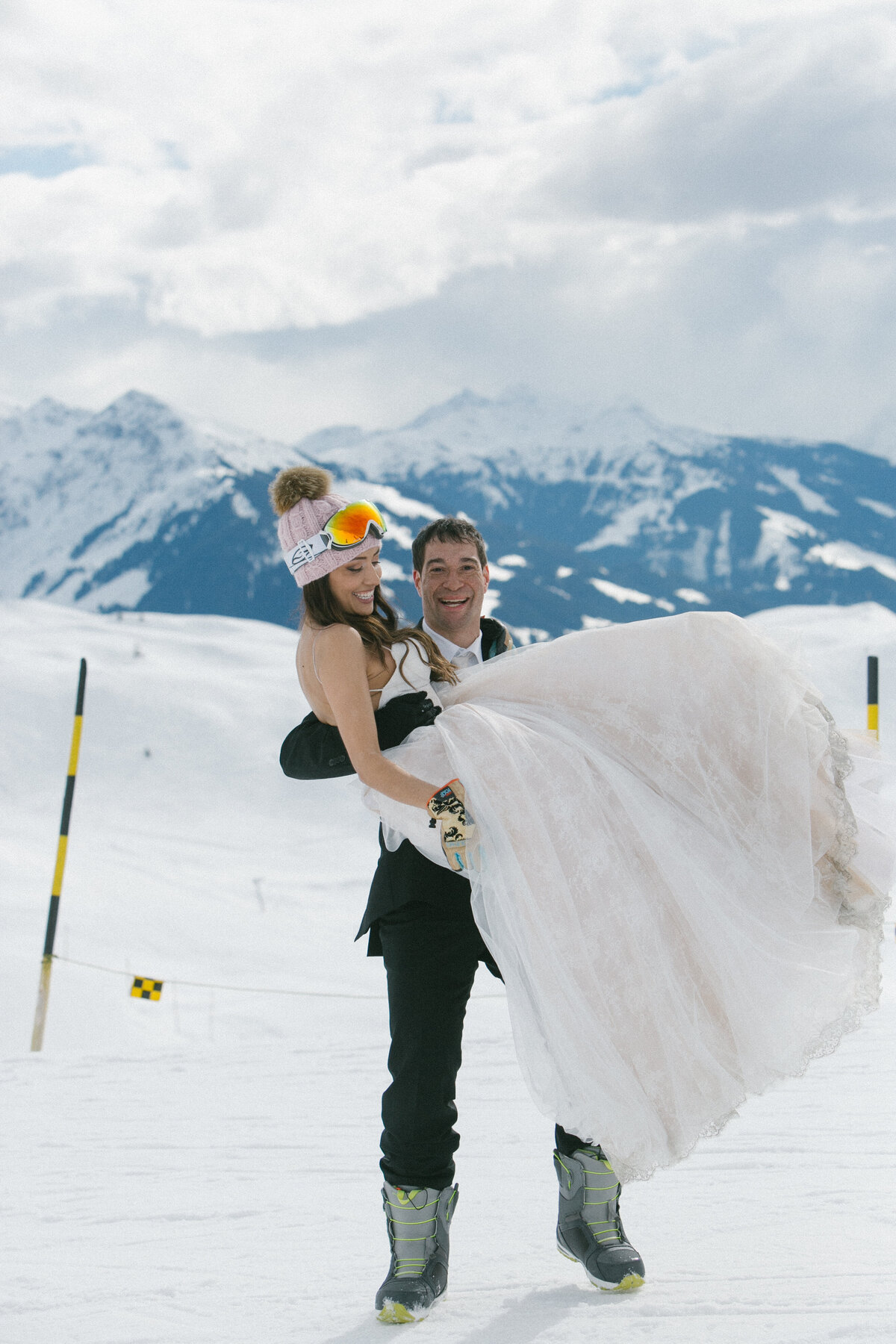 Bride, groom, and friends laughing in snow with snowboards and goggles – après-wedding energy in Kitzbühel by Studio Rose