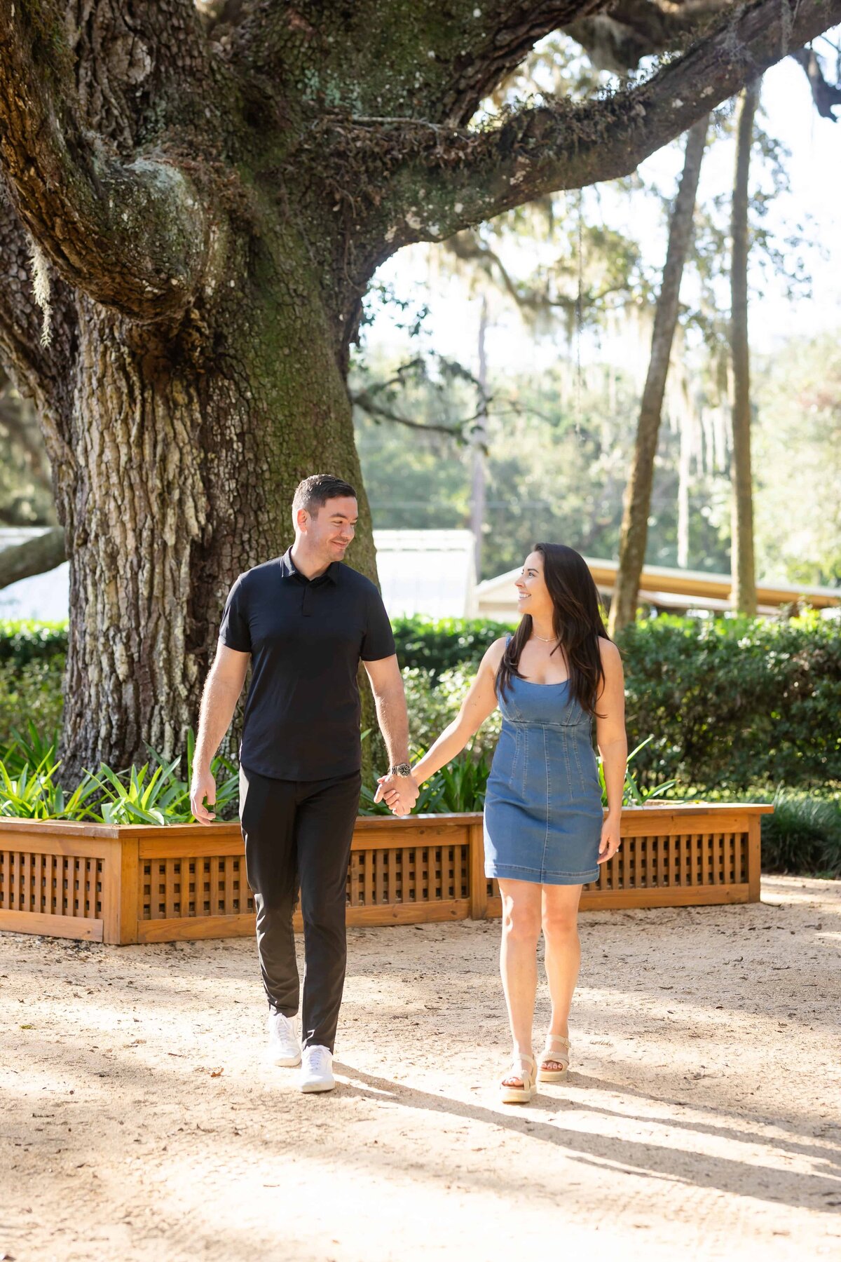 A couple walks hand-in-hand through a sunlit park in Jacksonville, Florida, during their engagement session. Surrounded by tall trees, Spanish moss, and soft natural light, the bride-to-be wears a denim dress and the groom-to-be sports a black polo and pants. Their relaxed smiles and connected stride reflect a comfortable, joyful love. Captured by Heartful Films, a Jacksonville engagement photographer and videographer specializing in natural, true-to-you storytelling.