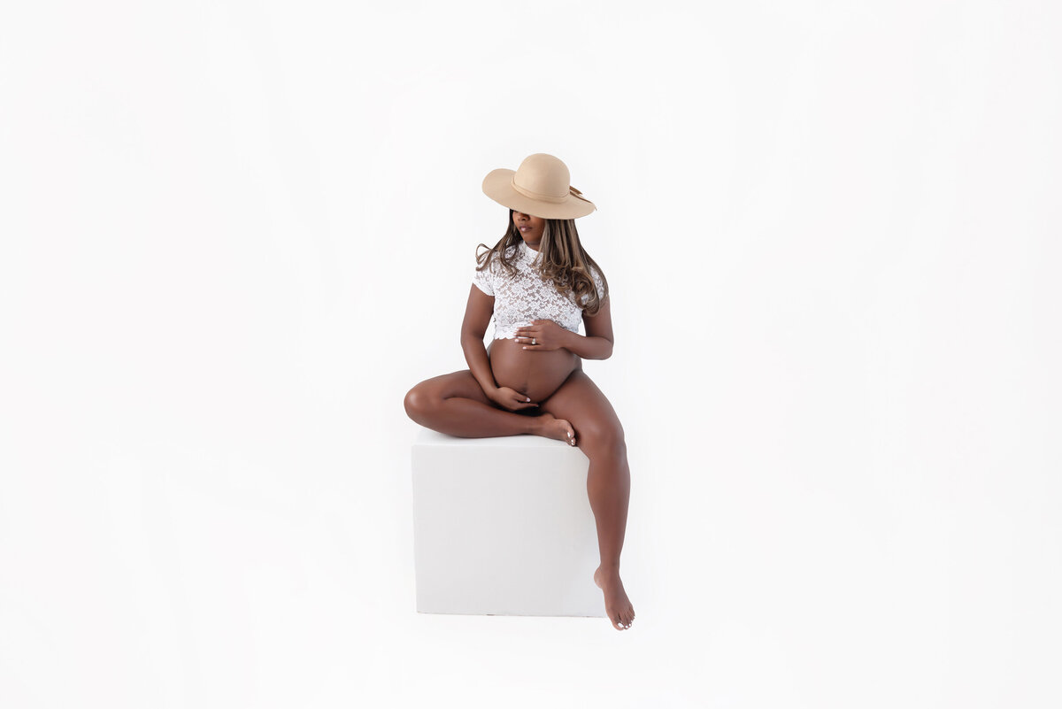 black pregnant woman wearing a white lace crop top sitting on a white block in front of a white background for her Burlington Studio Maternity Photography Session