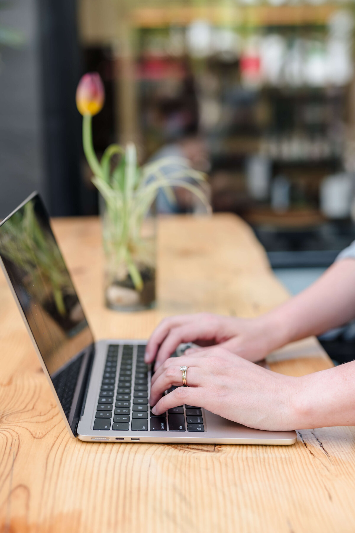 Close-up of hands typing on laptop at wooden table with blurred tulip in background.
