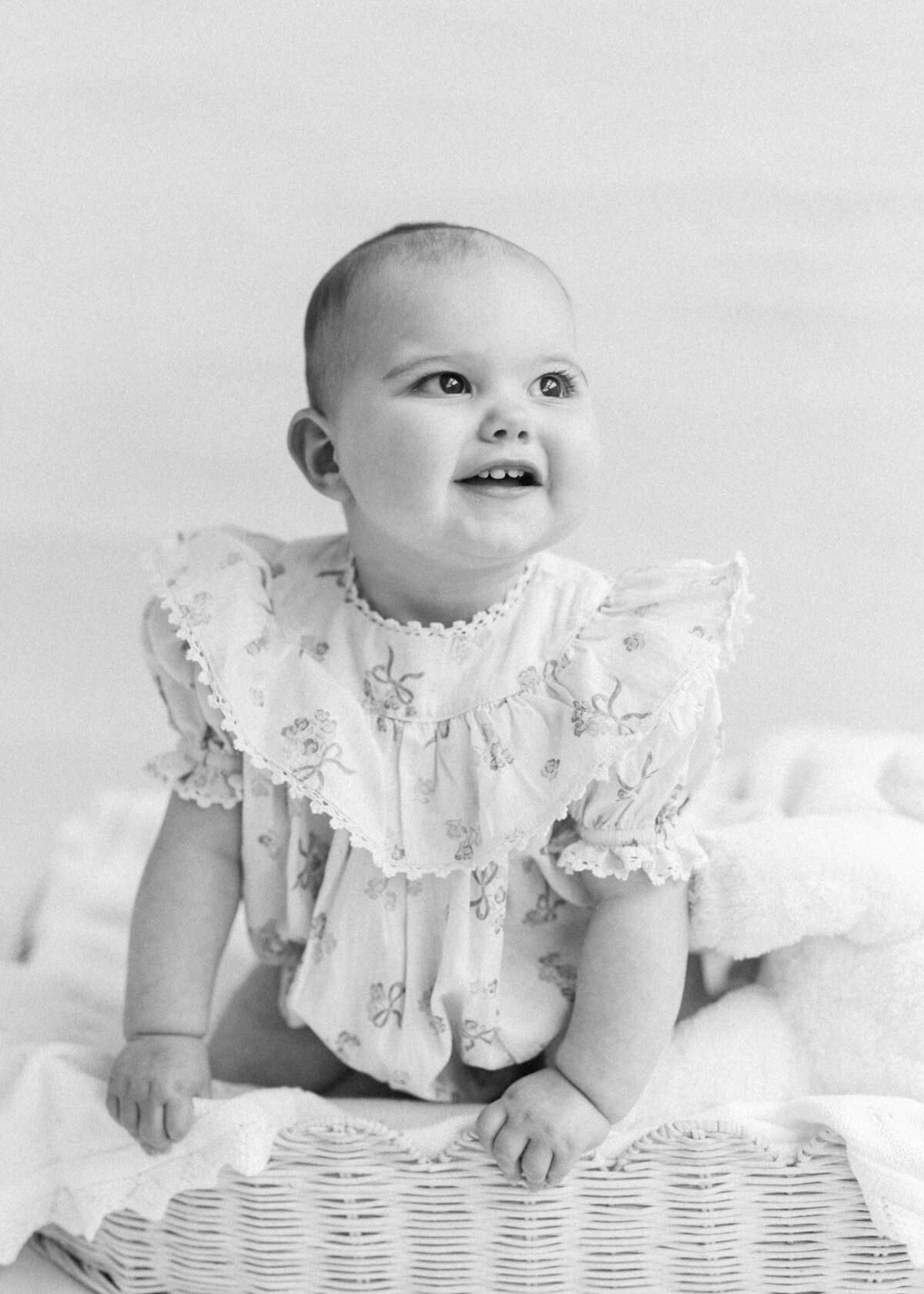 Black and white photo of a baby in a ruffly dress smiling away from the camera with her little teeth showing. 