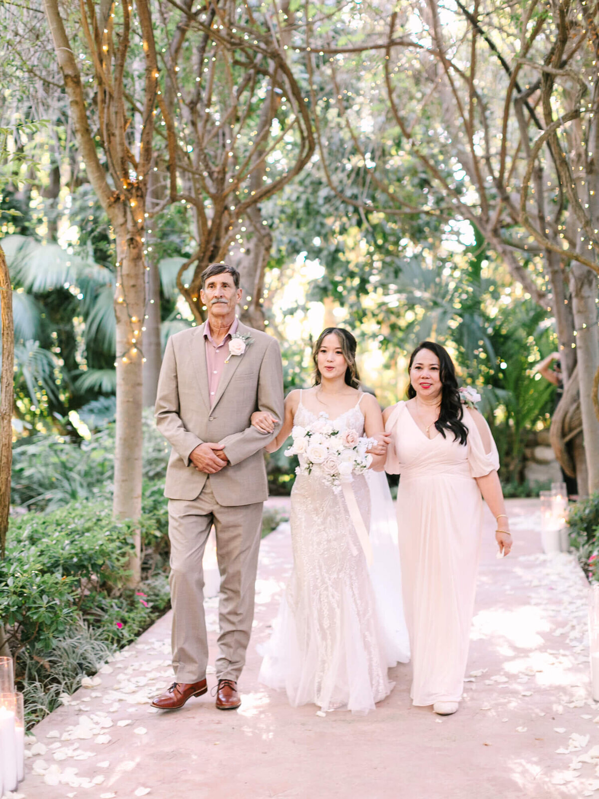 Bride walking down an outdoor aisle with dad and mom, surrounded by fairy-lit trees and white petals.