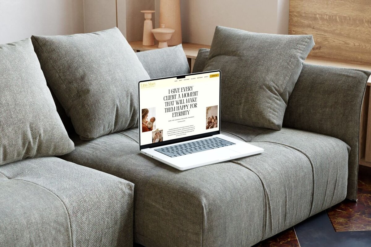 Open laptop placed on a gray cushioned sofa displaying a photographer’s website homepage with the headline “I give every client a moment that will make them happy for eternity” alongside warm-toned family portraits.