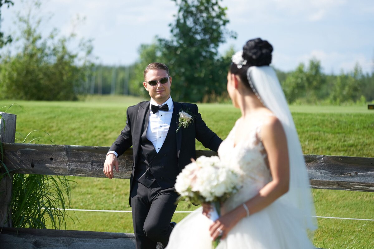 The bride glances over her shoulder with a warm smile at the groom, who is leaning casually on a fence. The moment captures a sense of playful affection and intimacy between the couple.