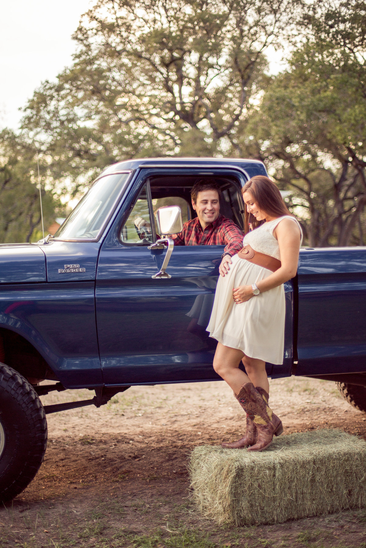 pregnant mother standing on a hay bail and father in s lifted vintage Ford truck for maternity session at park in San Antonio