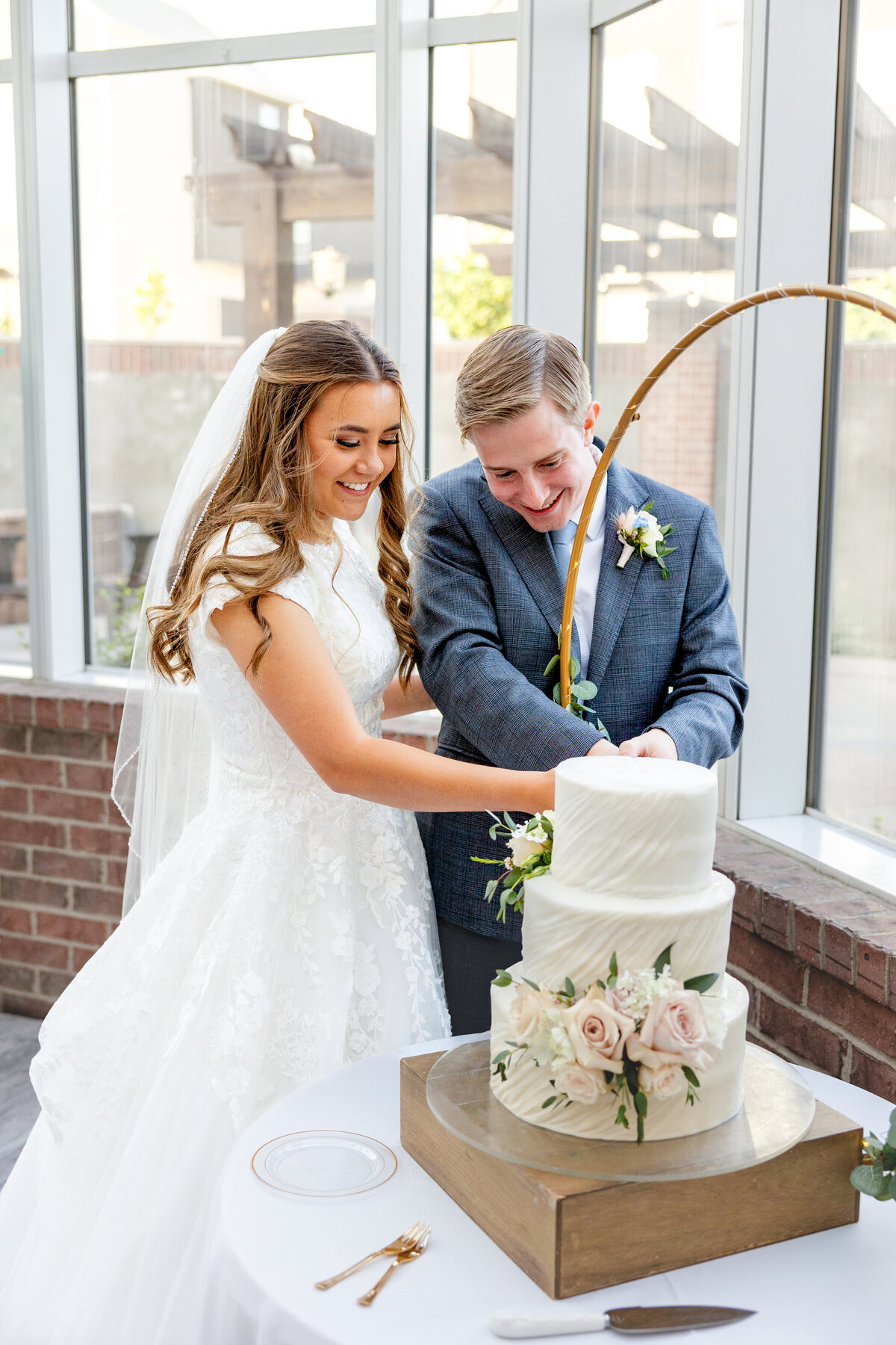 The bride and groom smile as they cut their beautifully decorated wedding cake together inside a sunlit reception hall.
