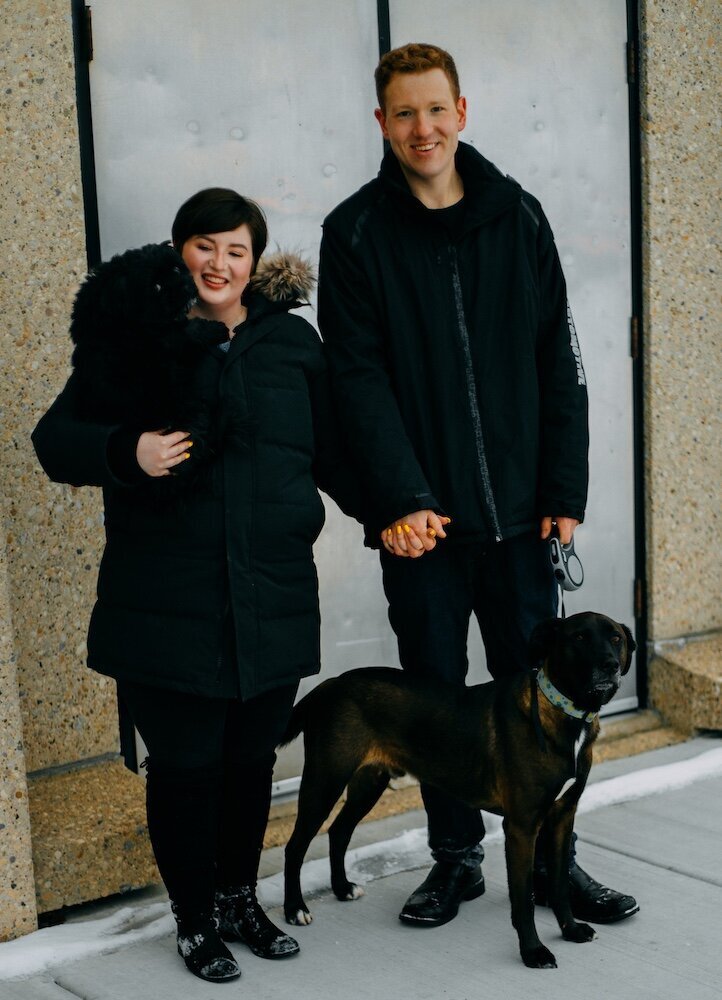 A couple stand holding hands while the woman on the left holds their little black dog in her arms and the man on the right has their bigger dog on a leash