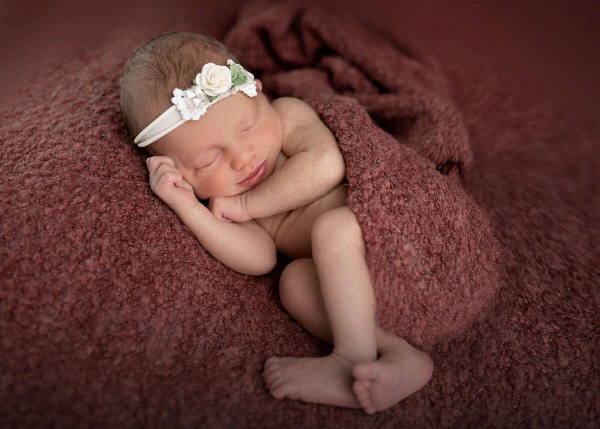 A sleeping newborn baby sleeps on her side in a white floral headband and maroon blanket