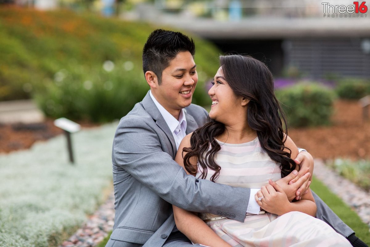 Groom to be embraces his fiance as she sits on his lap and looks back at him