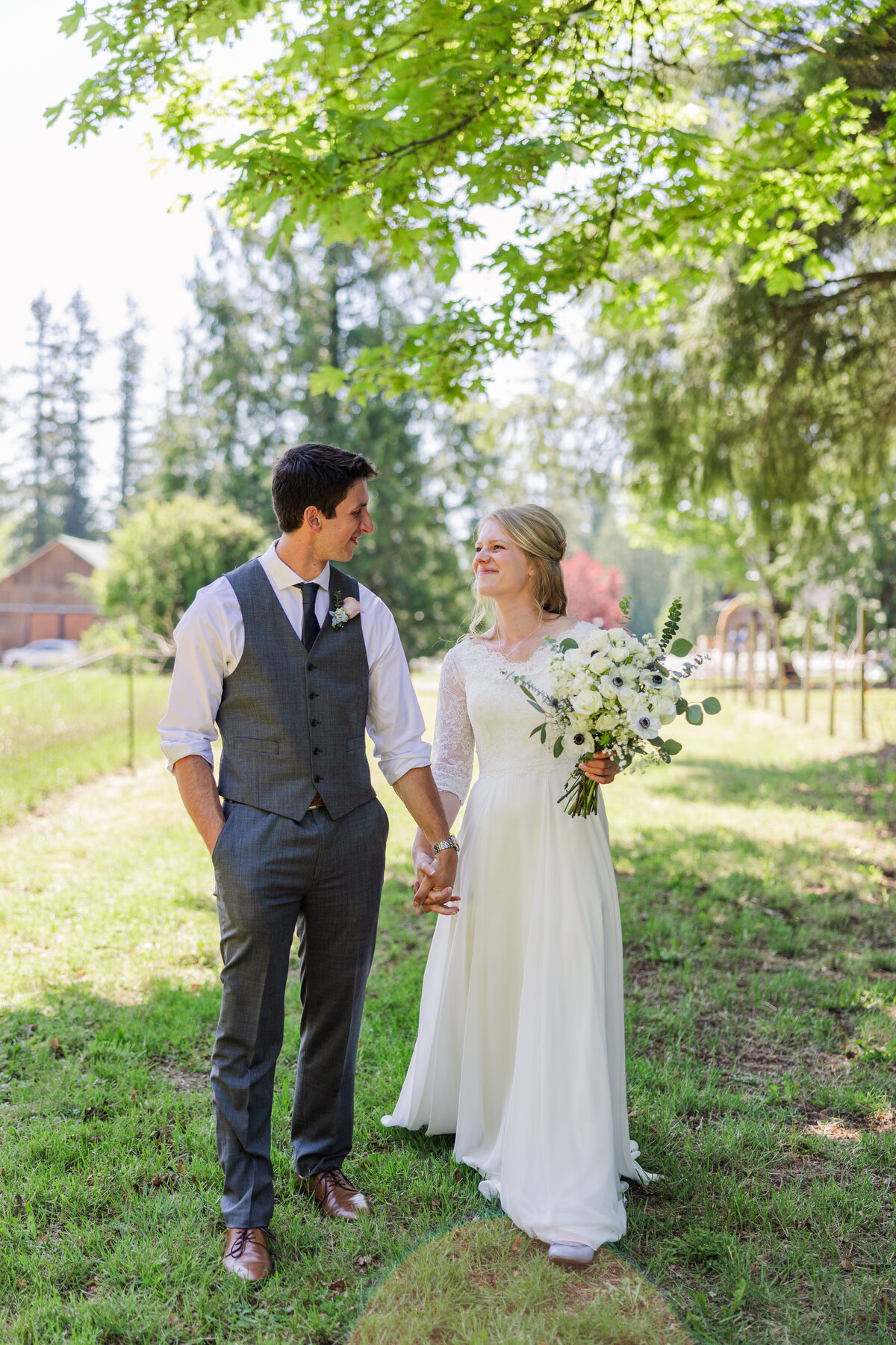 Bride & groom walk holding hands along a path of green trees and lush grass, during their wedding day couple photos
