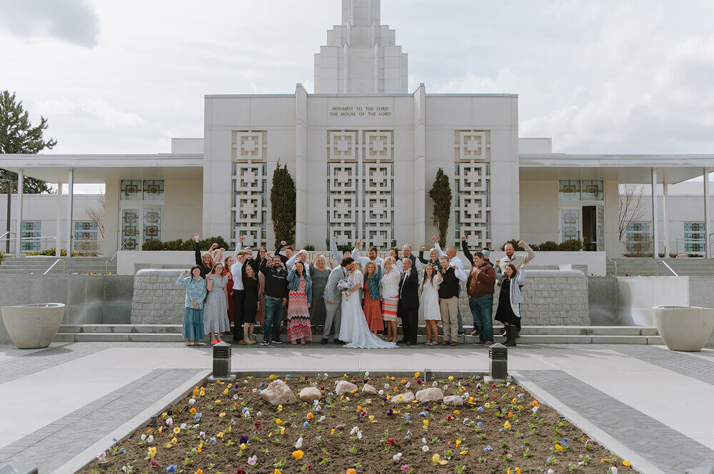 A bride and groom kiss in front of an LDS temple on their wedding day in Boise, ID.  Next to them are their friends and family celebrating.