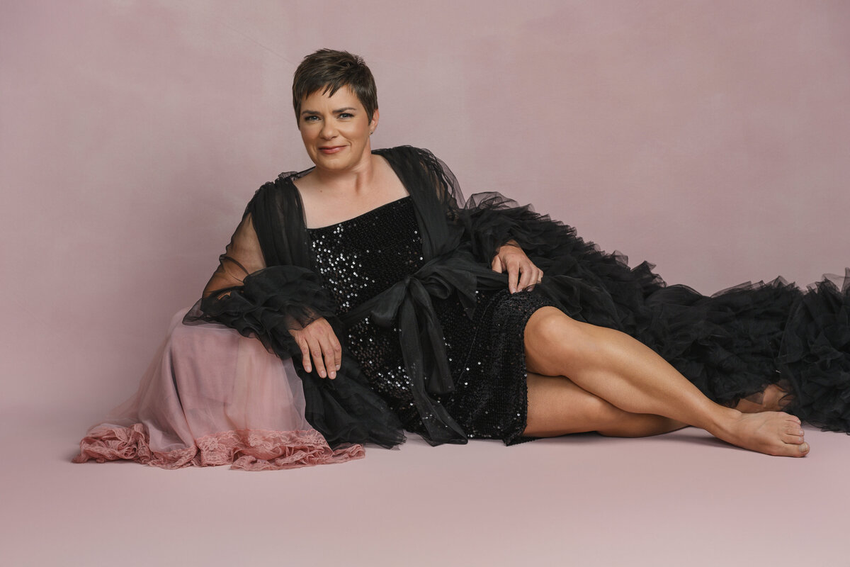 Woman in a black sequin dress with a dramatic tulle cape, reclining on a pink backdrop