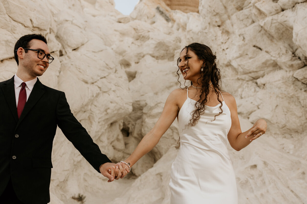 bride and groom walking while holding hands in front of a white rock