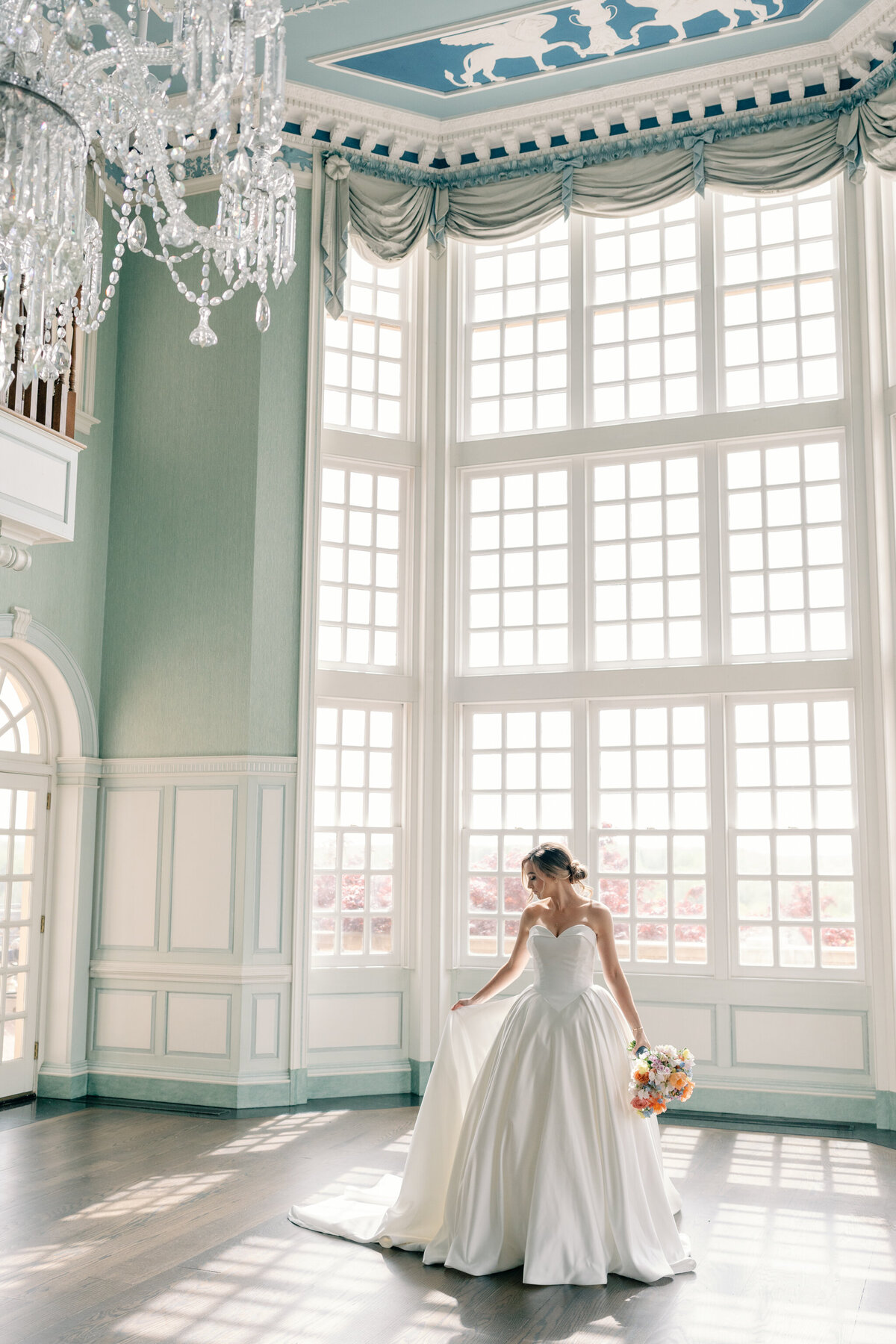 Wedding at the Estate at River Run in Maidens, VA | Wedding Photographer in Richmond | bride holding the train of her wedding dress and a colorful wedding bouquet in front of the large window in the ballroom of the Estate at River Run in Richmond
