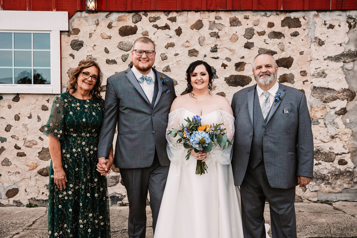Newlyweds hold hands and smile with mom and dad on either side in front of a stone wall