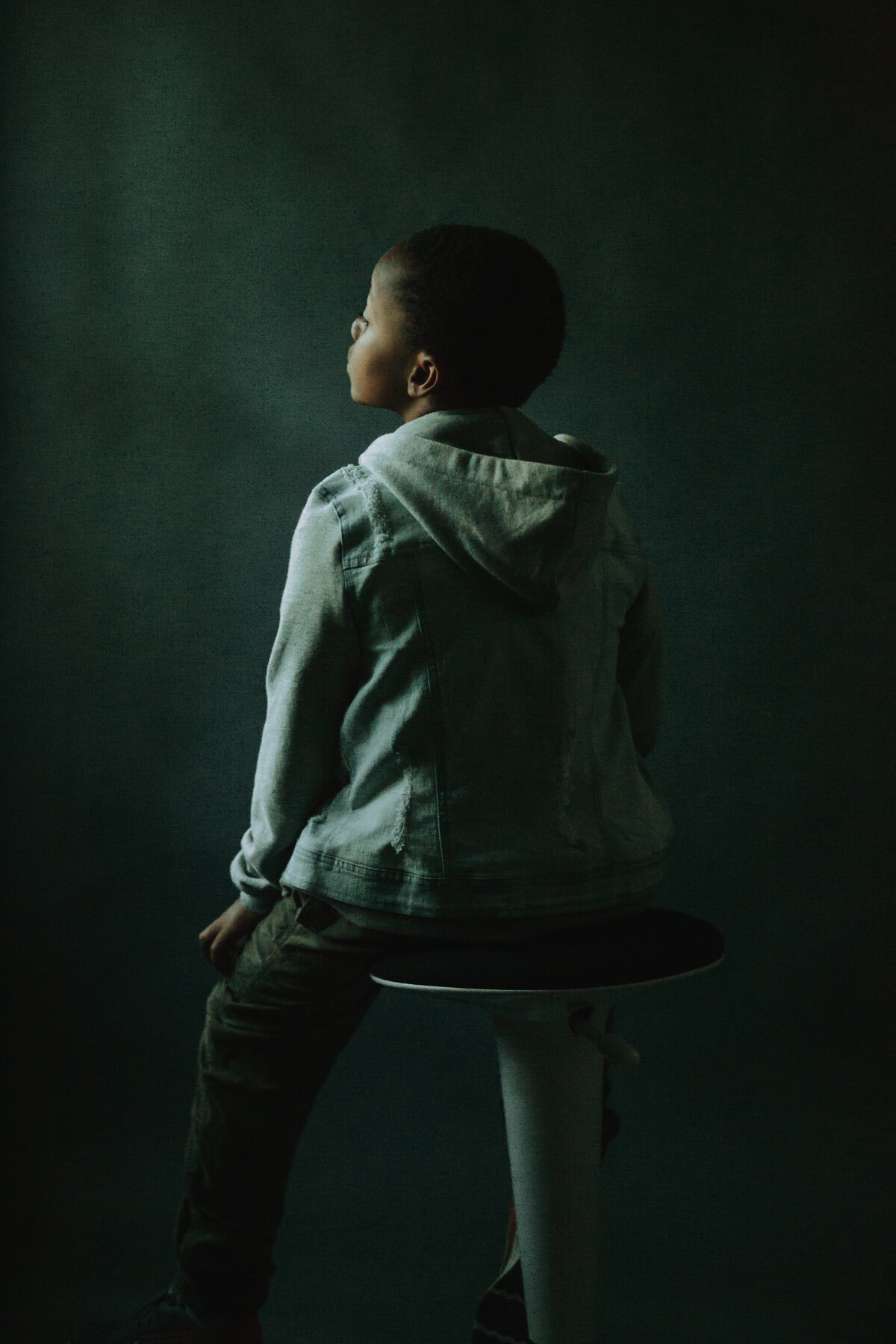 Picture of a boy sitting on a stool, facing away from the camera. Houston Fine Art Portraits.