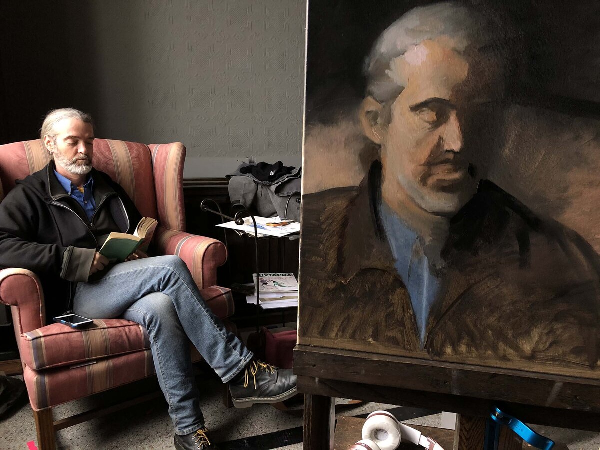 Oil painting of man with a grey beard reading a book with real subject posing beside it