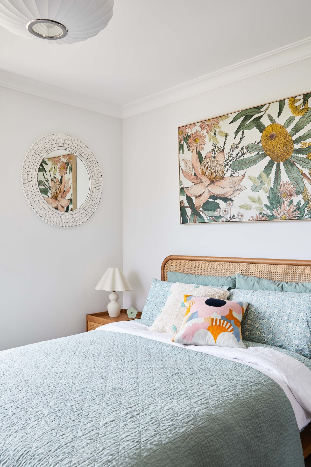 A light-filled bedroom interior featuring a bed with a rattan headboard and colorful pillows. A large botanical art piece hangs above the bed, and a round white wicker mirror is on the adjacent wall.