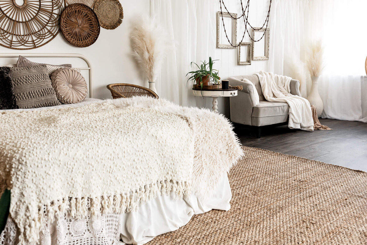 A cozy bedroom with a bed featuring textured blankets and pillows, a woven wall decor, and a soft area rug. A small round table with plants sits next to an armchair draped with a blanket, all against a backdrop of sheer white curtains.