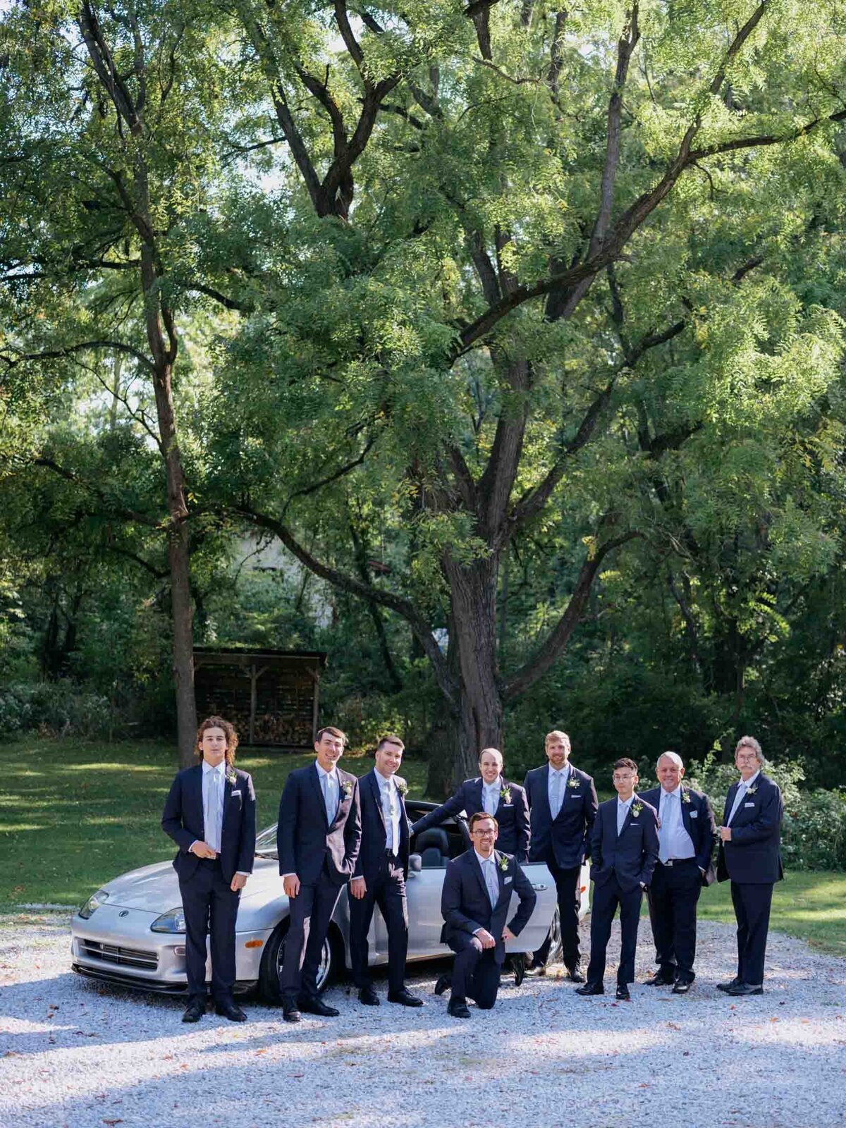 Groom in sports car surrounded by groomsmen.