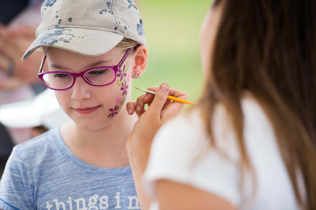 a face paint artist painting pink flowers on a little girl's face at a corporate children's event.  Captured by Ottawa Event Photographer JEMMAN Photography COMMERCIAL