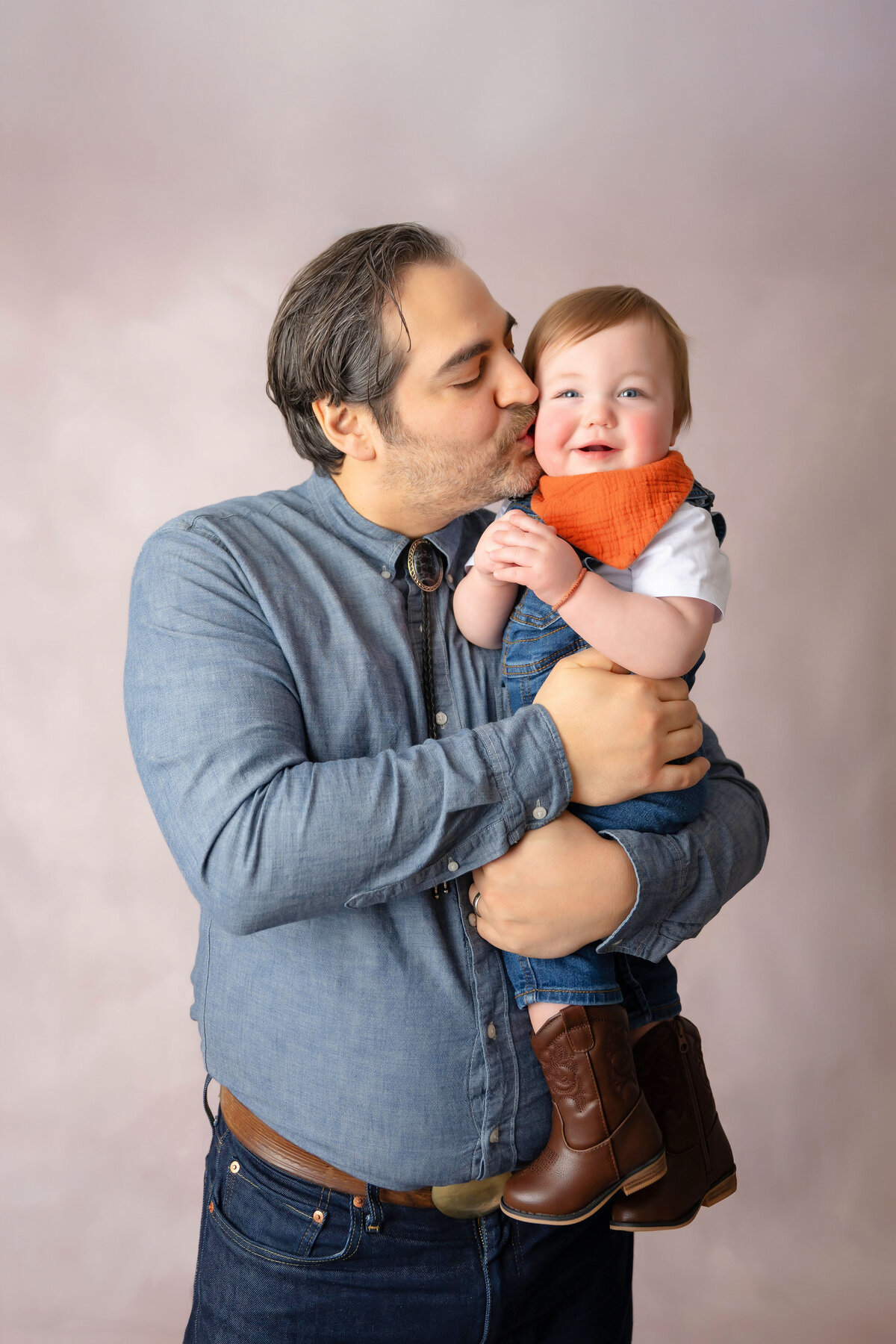 Dressed in matching jean clothing,  dad is holding baby while kissing him on the cheek. 