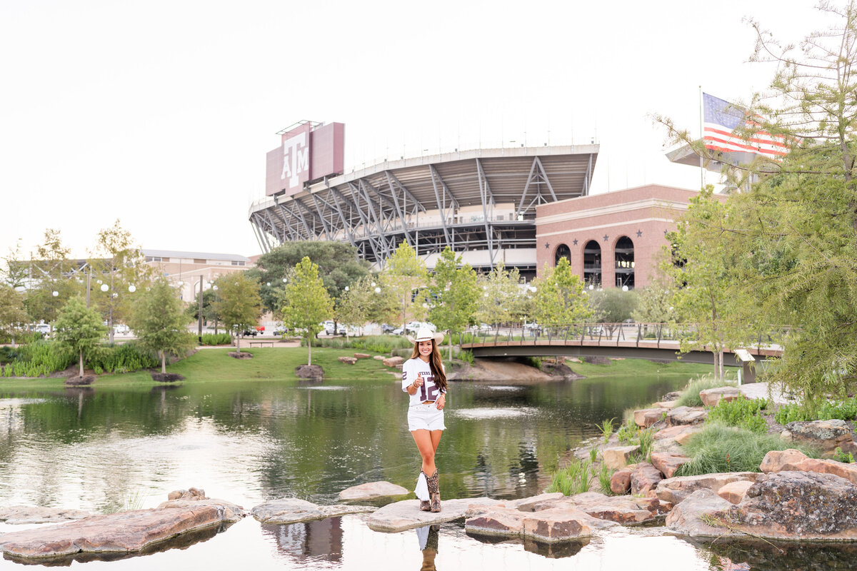 Texas A&M senior girl with hand on hip and thumbs up sign while standing on rocks with Kyle Field in background in Aggie Park while wearing all white jersey, shorts and cowboy hat