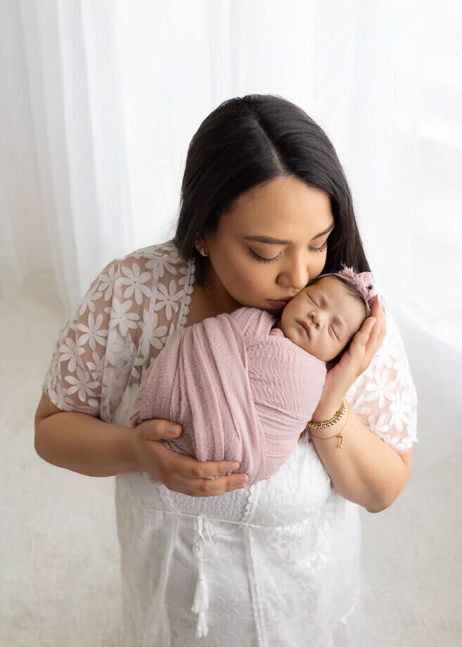 Mother in white holding her newborn girl in front of sheer white curtains