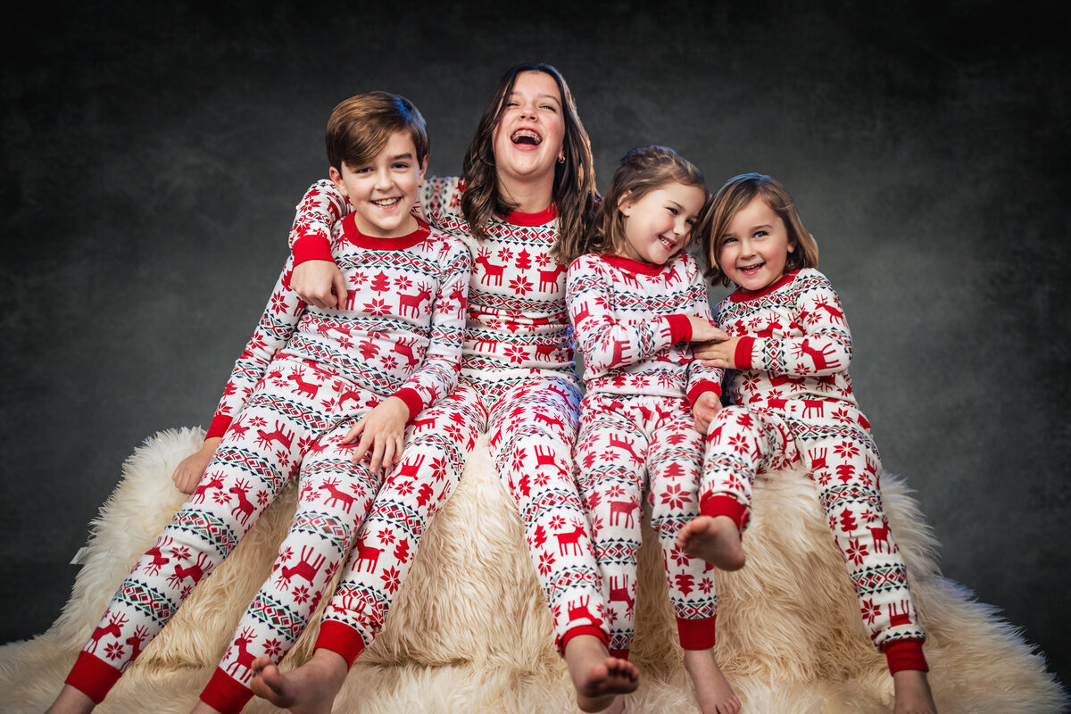 Four siblings embrace in Christmas pajamas on a white ottoman during Christmas mini sessions at the Pittsburgh photography studio they are all laughing and tickling each other