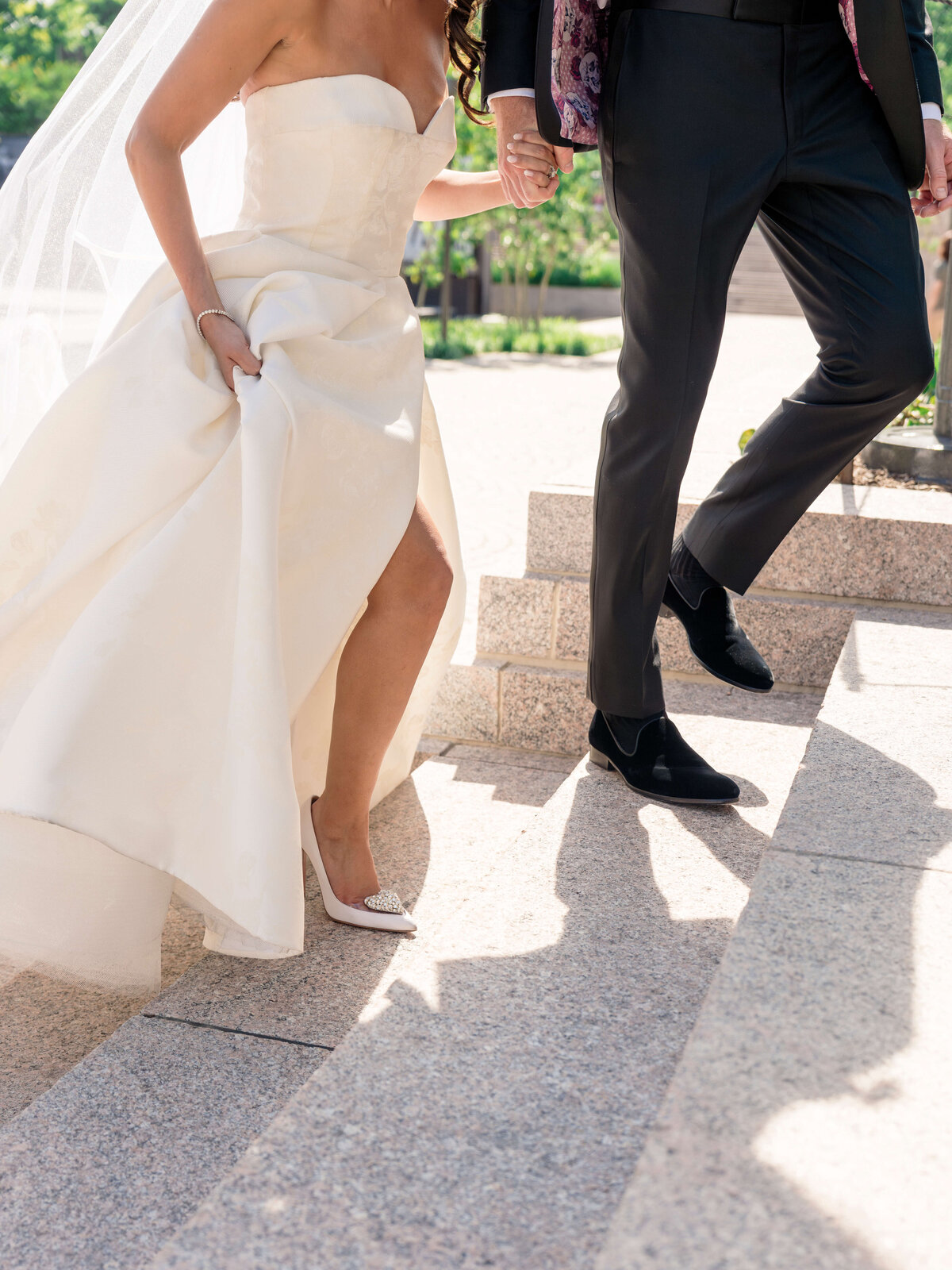 A bride in a strapless white gown walks up stone steps, holding her dress with one hand. Beside her is a groom in a dark suit, holding her hand. Sunlight casts shadows on the steps, and greenery is visible in the background.
