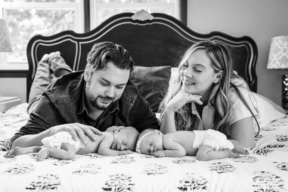 A black and white photo of a couple lying on a bed, lovingly gazing at their newborn twins who are lying between them.