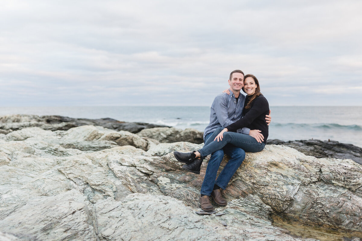 Engaged couple cuddle at Brenton Point Park with the sunset in the distance