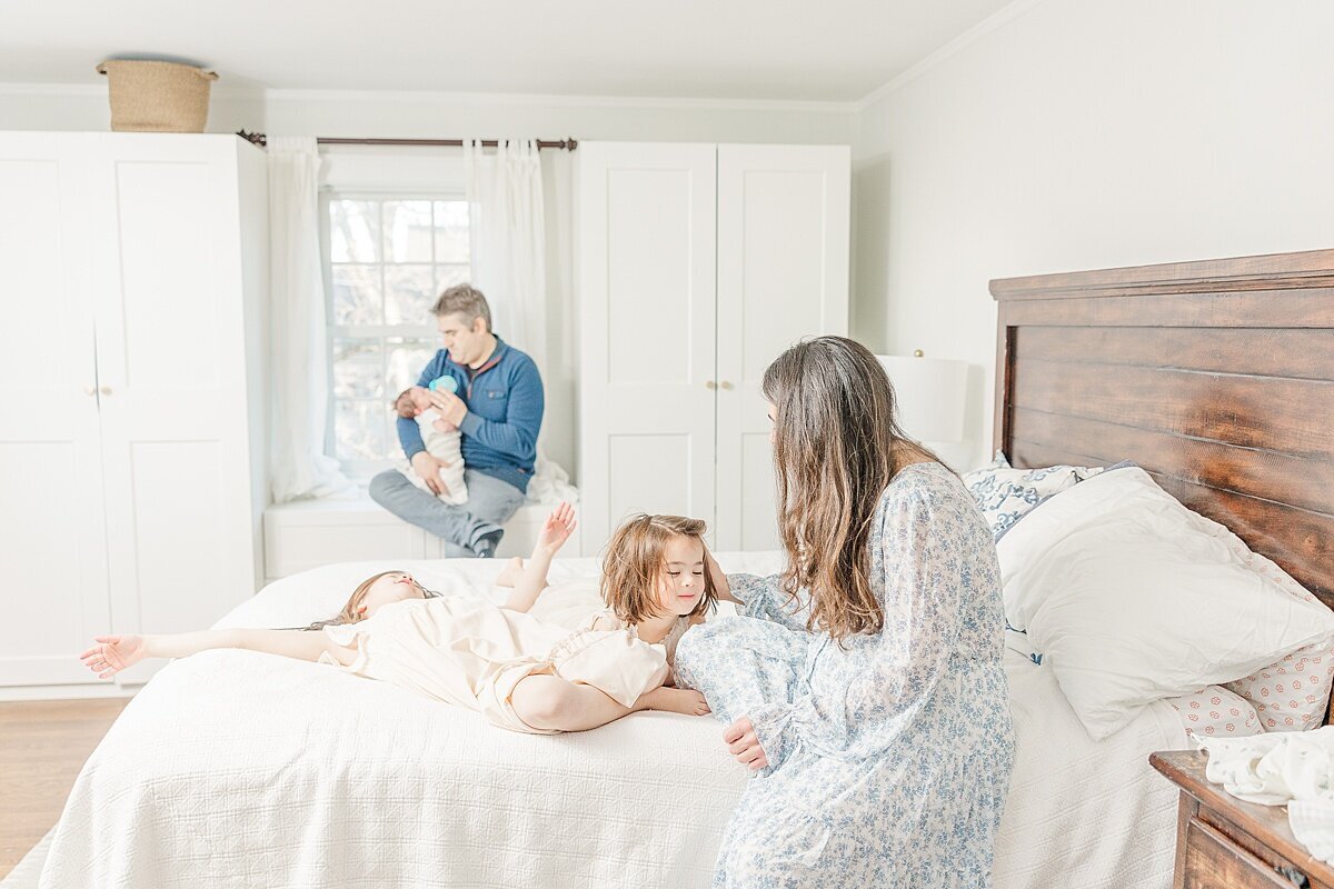 sisters play on bed while dad sits with baby dung in-home newborn photo session with Sara Sniderman Photography in Needham Massachusetts