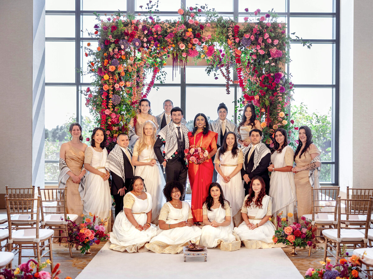 A wedding ceremony with a diverse bridal party. The couple stands under a floral arch, surrounded by attendants wearing coordinated attire. The setting is bright and elegant, featuring colorful flowers and natural light streaming through large windows.