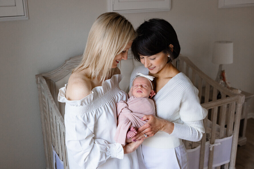 Two moms in white hold and look at their new baby on the baby's nursery.