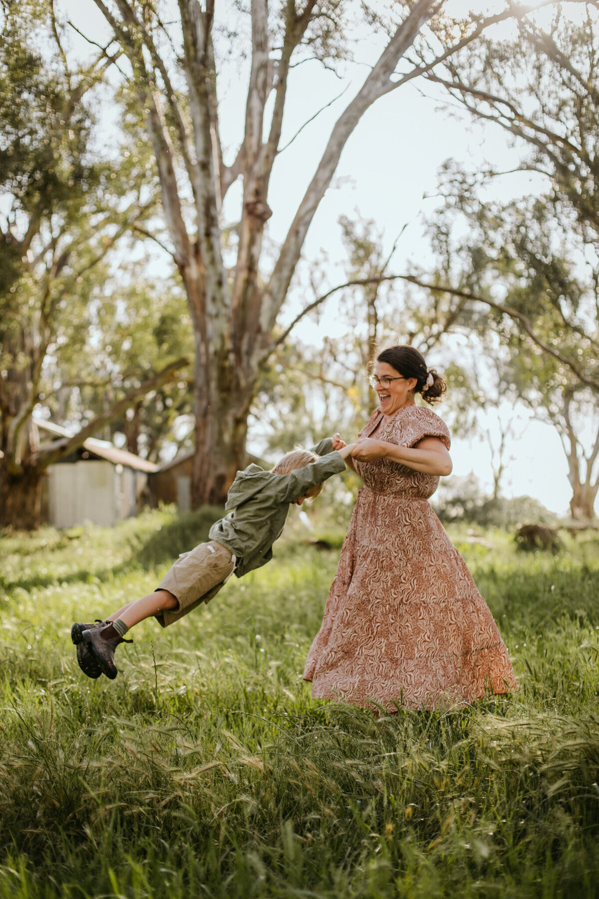 Child laughs as mom swings him by the arms in a sunlit grass field.