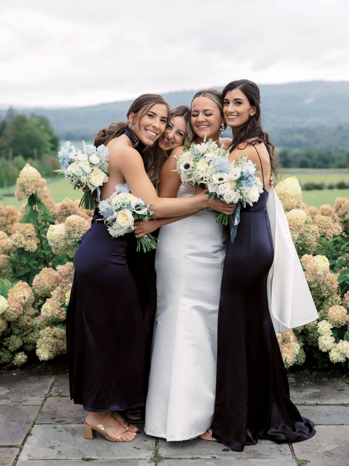 The bride and her bridesmaids are happily hugging each other in a garden at The Lion Rock Farm, CT. Image by Jenny Fu Studio