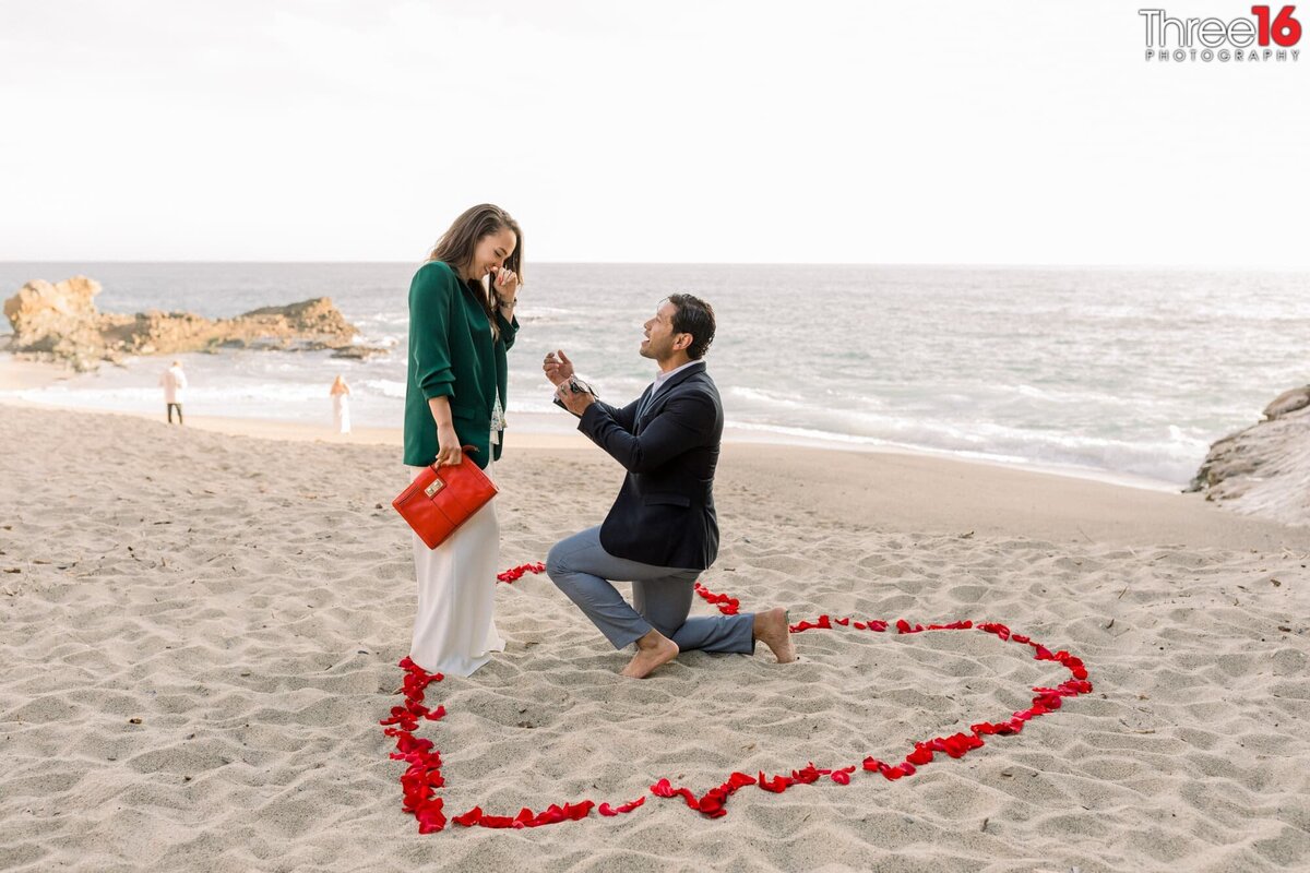 Woman is surprised as her boyfriend is on one knee to propose marriage while in a heart shaped out of red roses on the beach