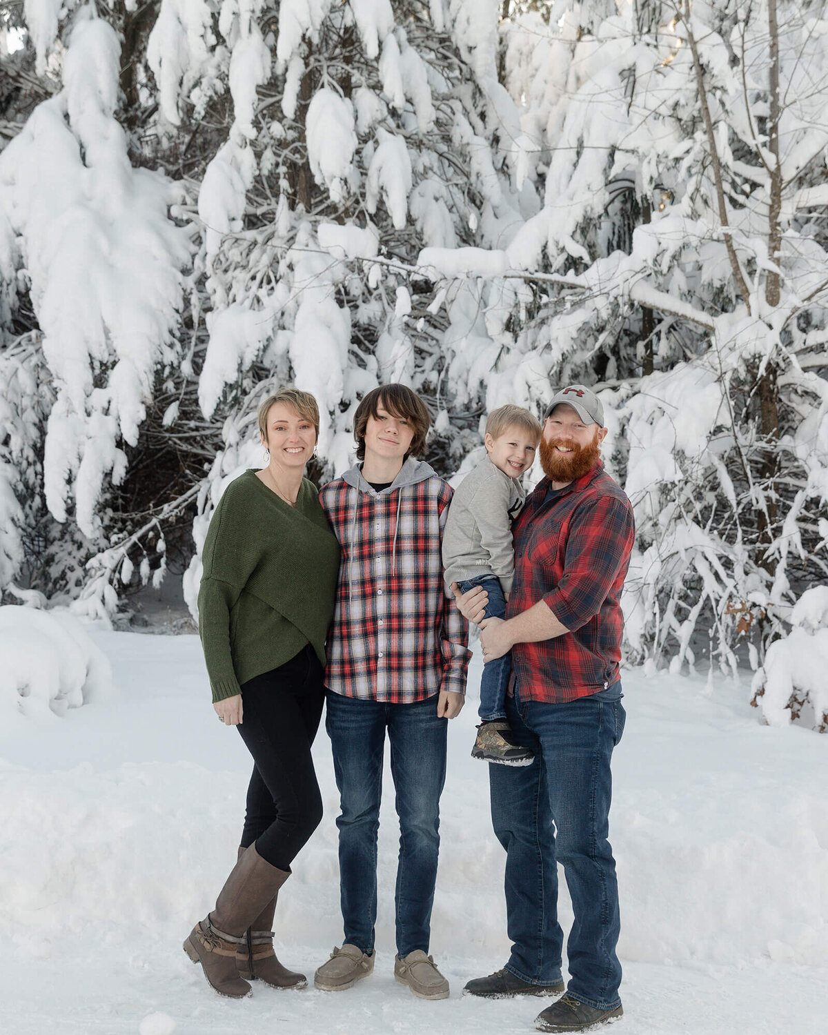 Family of 4 standing outside in front of trees with heavy snow on them. 